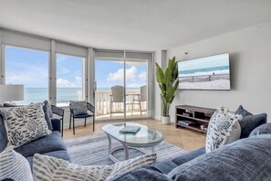 A living room with a glass coffee table, a sofa, and a TV on the wall. There is a balcony with a view of the beach and ocean. - New Listing! Updated Oceanfront 2/2 | Heated Pool & Garage Parking (Ormond Beach)