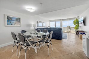 Een moderne woonkamer met een eethoek, een bank, een televisie en een schilderij van een strandgezicht.