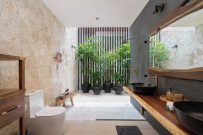 Een moderne badkamer met een stenen muur, een houten wastafelmeubel, een grote zwarte wasbak en diverse potplanten.