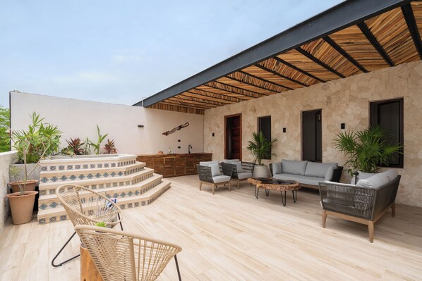 Een overdekt terras met een stenen muur, een houten trap, rieten meubels en potplanten.