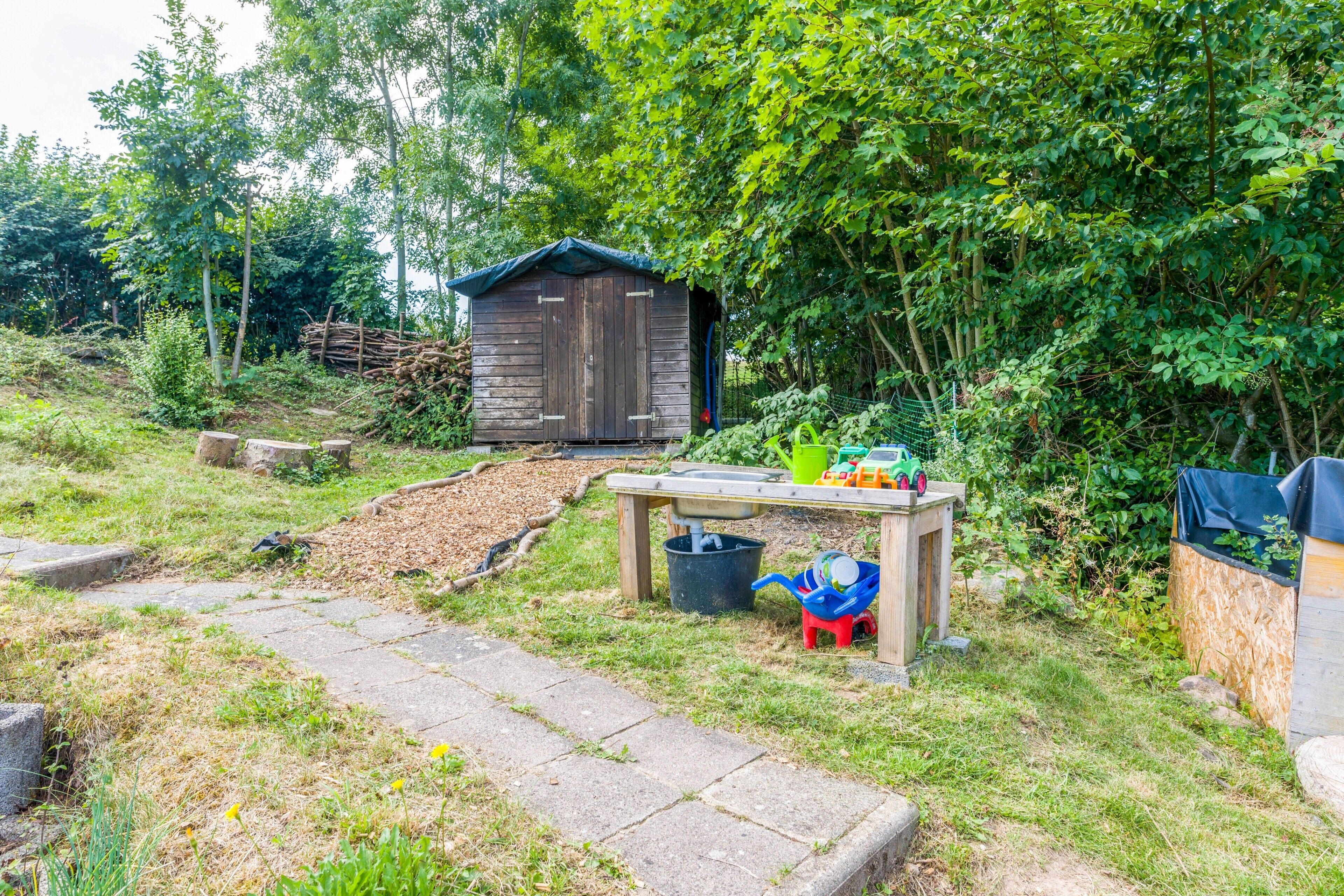 A wooden shed with a sloped roof, a small table with a sink, and a garden area with a wooden path.