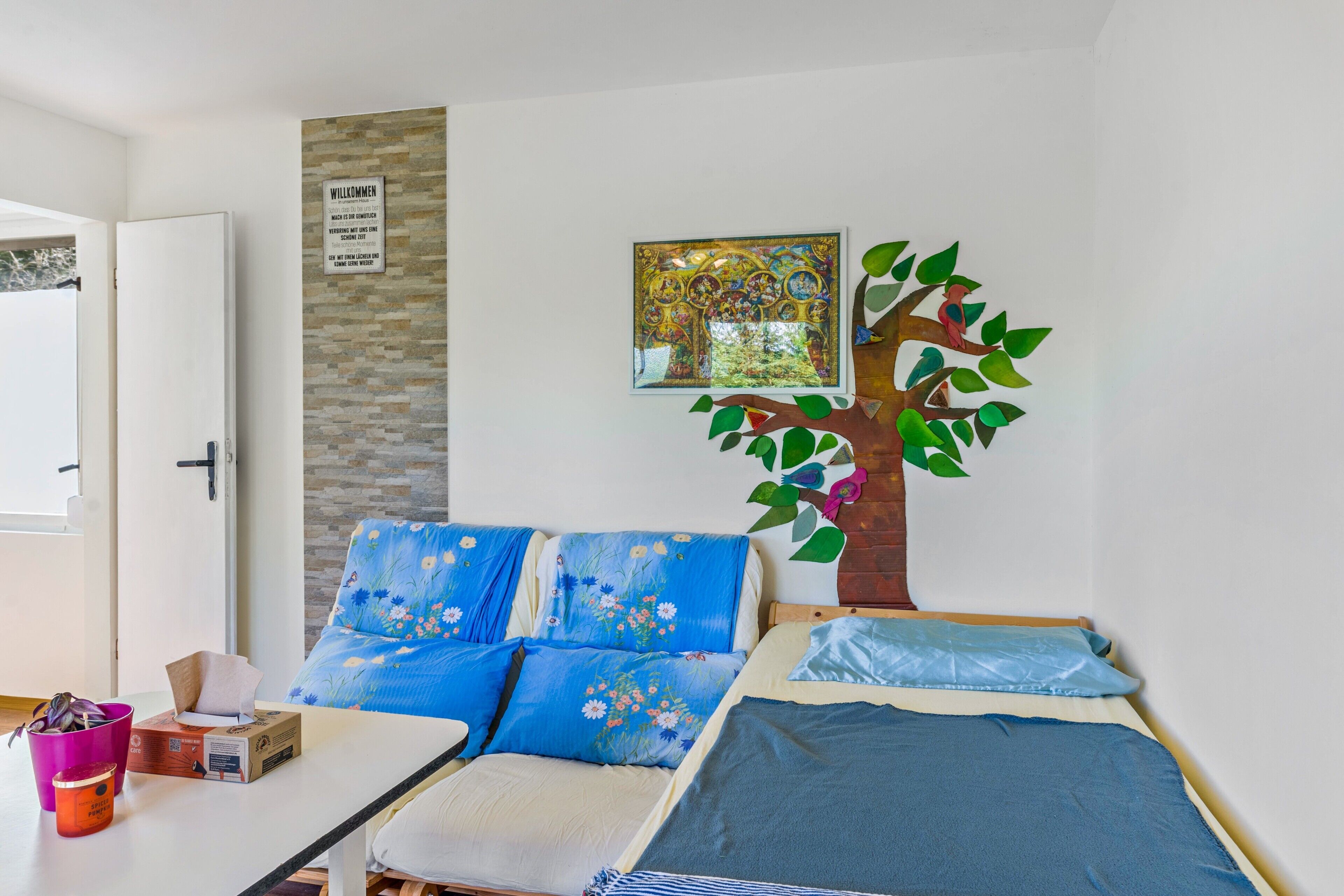 A bedroom with a bed, a sofa, a wall-mounted tree decoration, a welcome sign, and a window.