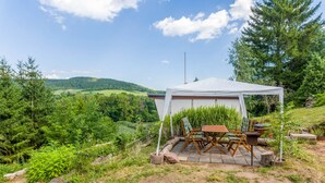 A tented outdoor seating area with a table and chairs, surrounded by greenery and hills in the background.
