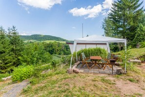A tented outdoor seating area with a table and chairs, surrounded by greenery and hills in the background.