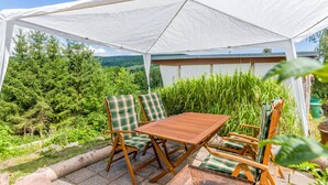A covered outdoor seating area with a table and chairs, surrounded by greenery and a view of the landscape.