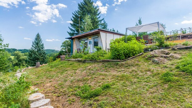 A small cabin with a sloped roof, surrounded by lush greenery and trees.