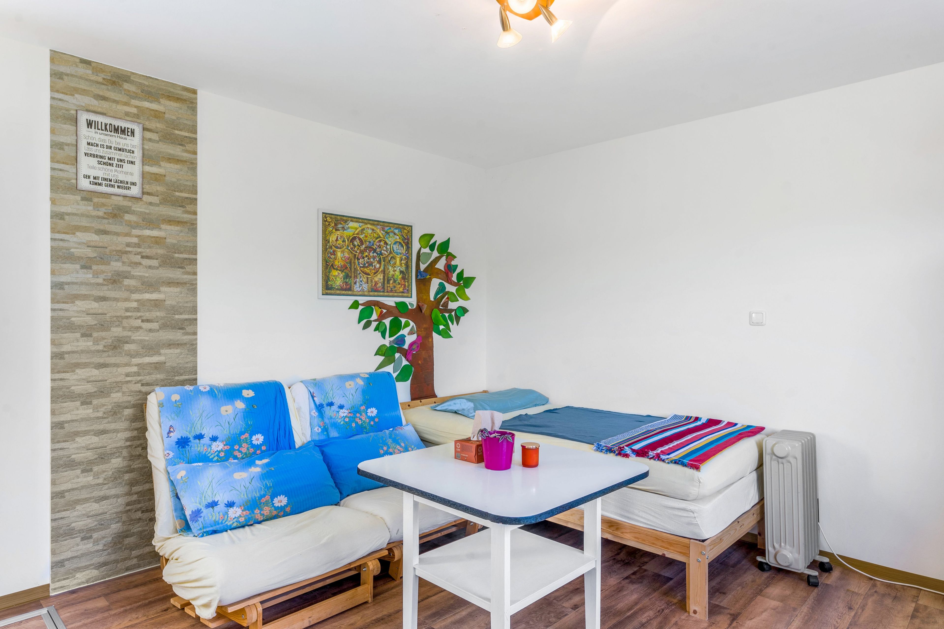 Una habitación pequeña y acogedora con un sofá, una mesita y una cama. Hay un cartel de bienvenida en la pared, un árbol decorativo y un radiador.