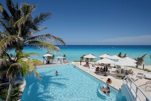 A pool area with clear blue water, surrounded by lounge chairs and umbrellas, with a view of the ocean and palm trees. - Beachfront studio / Amazing Caribbean view (Cancún)