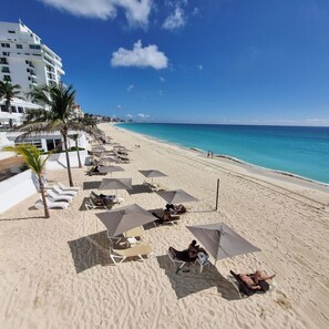 A beach with sun loungers and umbrellas, a clear sky, and a building in the background. - Beachfront studio / Amazing Caribbean view (Cancún)