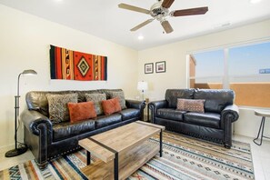 Ein Wohnzimmer mit zwei Ledersofas, einem Couchtisch, einem gemusterten Teppich, einem Deckenventilator und einem großen Fenster.
