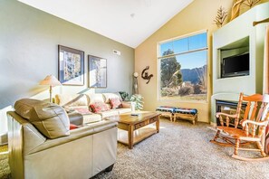 A living room with a sofa, coffee table, and a rocking chair. There is a television mounted on the wall, and a window with a view of the outdoors. - Colorful Condo with Shared Pool and Hot Tub - Close to Golf & Arches National Park (Moab)