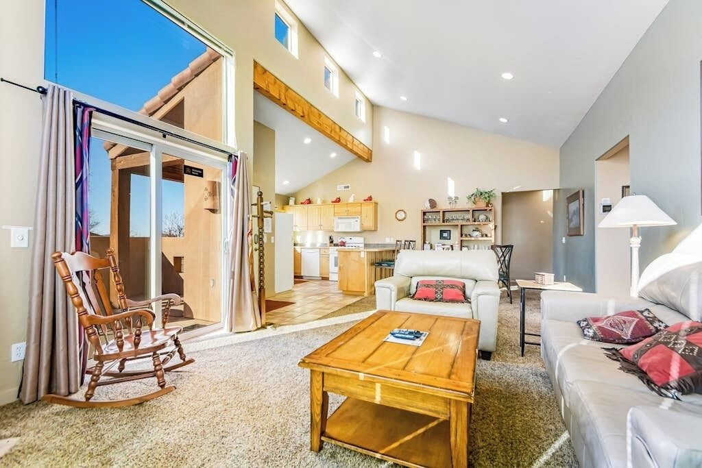A spacious living area with a sofa, a coffee table, and a rocking chair. There is a kitchen in the background with a dining table and chairs.