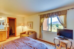 Ein Schlafzimmer mit einem Bett, einem Fernseher, einem Schreibtisch, einem Stuhl, einem Fenster mit Vorhängen und einer Tür zu einem anderen Raum.