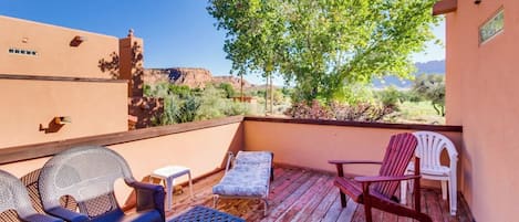 Una terraza de madera con mobiliario exterior, vista a un árbol y un edificio al fondo.
