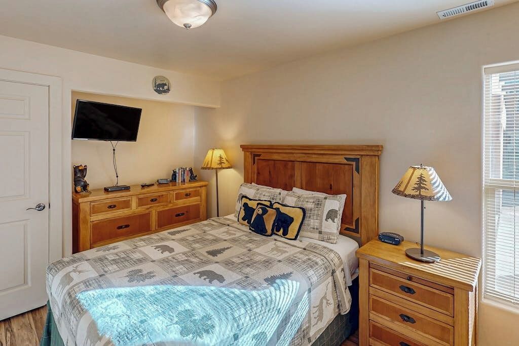 Un dormitorio con una cama, un televisor montado en la pared, un aparador de madera y dos lámparas de noche.