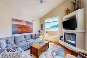 A living room with a sofa, coffee table, fireplace, TV, and a large painting. - Spacious Moab Townhouse with Seasonal Pool and Hot Tub - Views and Hiking Nearby (Moab)