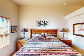 A bedroom with a wooden bed, patterned bedding, two bedside lamps, a wall-mounted artwork, and a window with blinds. - Spacious Moab Townhouse with Seasonal Pool and Hot Tub - Views and Hiking Nearby (Moab)