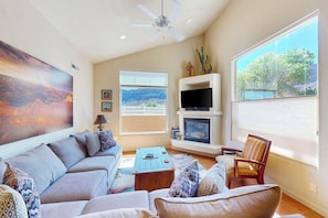 A living room with a sofa, coffee table, TV, fireplace, and large windows with a view. - Spacious Moab Townhouse with Seasonal Pool and Hot Tub - Views and Hiking Nearby (Moab)