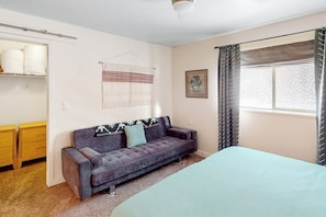 A bedroom with a bed, a sofa, a window with curtains, a wall-mounted shelf with towels, and a framed picture. - Spacious Moab Condo with Pool Hot Tub and Mountain Views (Moab)