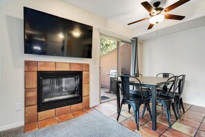 A living room with a fireplace, a flat-screen TV mounted above it, a dining table with chairs, and a ceiling fan. - Remodeled Townhouse with Pool, Private Hot tub & Fireplace - Near National Park (Moab)