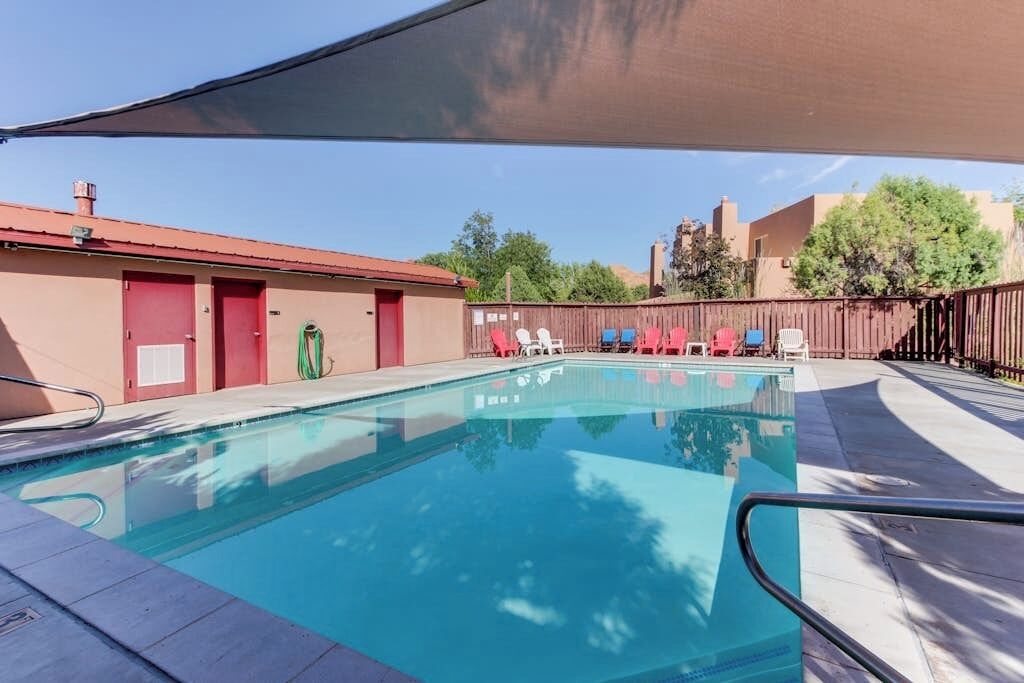 Una piscina con zona sombreada, una puerta roja y un patio trasero cercado con sillones de descanso.
