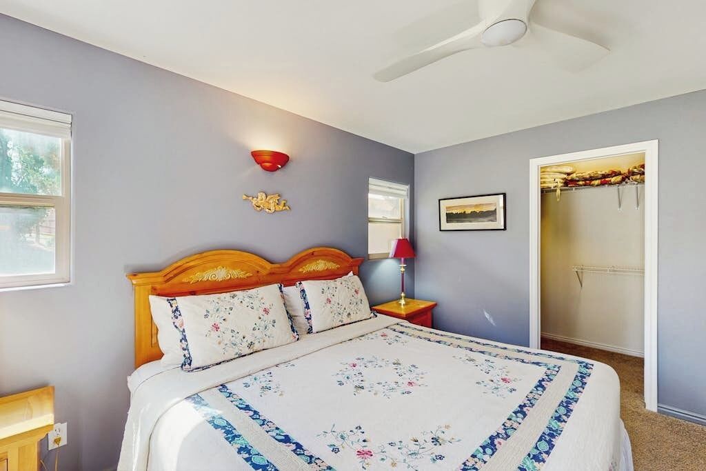 A bedroom with a bed, a nightstand, a wall-mounted lamp, a ceiling fan, and a door leading to another room.