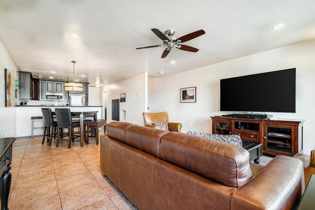 A modern living room with a leather sofa, a dining area with chairs, a kitchen with cabinets, a ceiling fan, and a flat-screen TV.