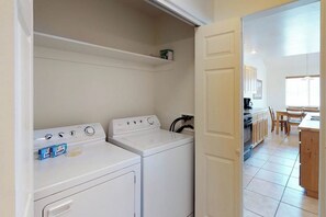 A laundry room with two white washing machines, a shelf above, and a kitchen area visible through an open door. - Spacious Townhome in a Great Location with Seasonal Shared Pool and Hot Tub (Moab)