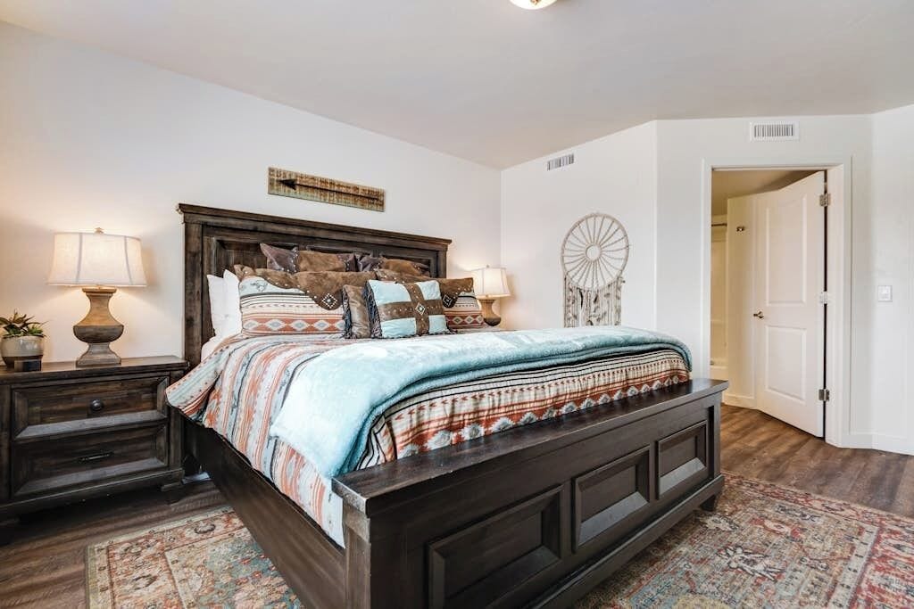 A bedroom with a dark wooden bed, patterned bedding, two bedside tables with lamps, a decorative wall piece, and a patterned rug.