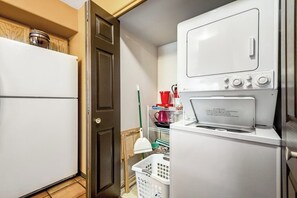 Uma lavanderia compacta com máquina de lavar e secadora, uma prateleira pequena com produtos de limpeza e uma porta que dá para outro cômodo.
