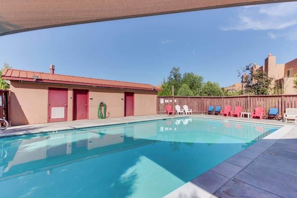 Une piscine avec un espace ombragé, un bâtiment avec des portes rouges et des sièges à l’extérieur.