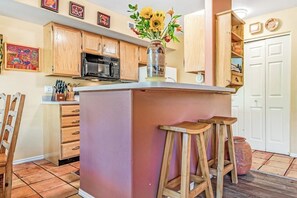 A kitchen with wooden cabinets, a central island with stools, a microwave, and a vase with flowers. - Condo Surrounded by Natural Red Rock Beauty with Seasonal Pool Access (Moab)