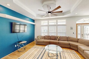 Una sala de estar con un sofá seccional, una mesa de centro de vidrio, un ventilador de techo y un televisor de pantalla plana montado en la pared.