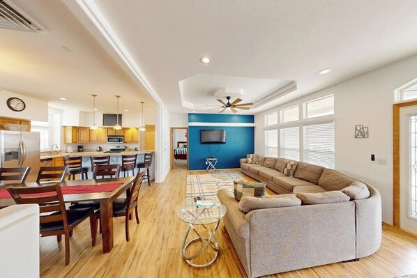 A modern living room with a sofa, dining table, and chairs. There is a kitchen in the background with cabinets and a refrigerator.