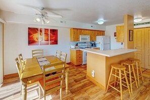 A compact kitchen with wooden cabinets, a central island with stools, and a dining area with a table and chairs. - Golf Course Front Condo with Seasonal Pool & Views - Walk to Steel Bender Trail (Moab)