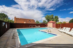 A swimming pool with a wooden deck, lounge chairs, and a building in the background. - Golf Course Front Condo with Seasonal Pool & Views - Walk to Steel Bender Trail (Moab)