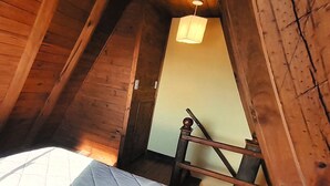 Huone, jossa on kalteva katto, puuseinät, peitolla varustettu sänky, riippuvalaisin ja puinen tuoli.