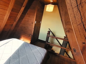 Una habitación con techo inclinado, paredes de madera, una cama con colcha, una lámpara colgante y una silla de madera.