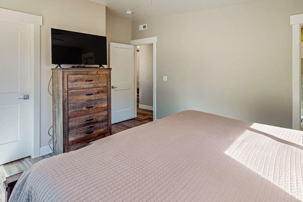 Un dormitorio con cama, cómoda de madera, televisor y puerta que da a otra habitación.