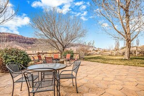 An outdoor patio with a round table and chairs, surrounded by stone flooring and sparse vegetation. - Dog Friendly Moab Mountain Escape with Provate Hot Tub, Fireplace, and Game Room (Moab)