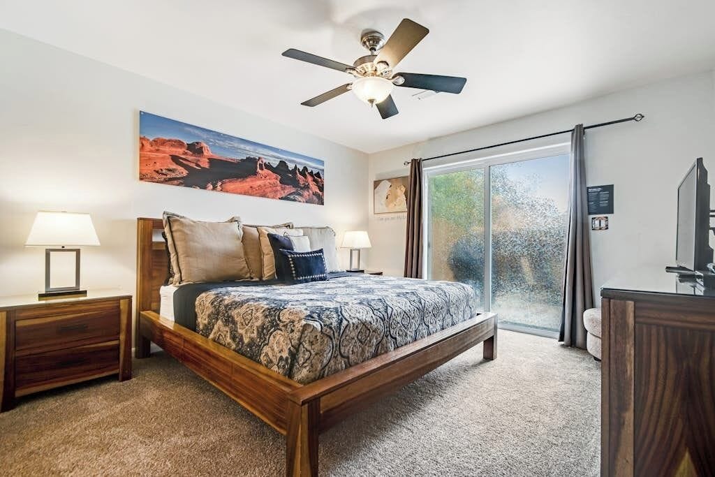 Een slaapkamer met een bed, een plafondventilator, een nachtkastje, een lamp, een raam met uitzicht en een deur.