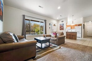 A living room with a sofa, a coffee table, and a view of the outdoors through a large window. - Updated Family Home with Private Hot Tub, Firepit & Shared Pool (Moab)