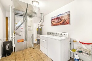 A laundry area with a washing machine, a dryer, and a water heater. There is a painting on the wall and a shelf with cleaning supplies. - Updated Family Home with Private Hot Tub, Firepit & Shared Pool (Moab)