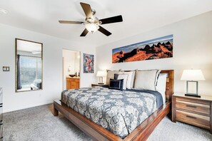 A bedroom with a bed, a ceiling fan, a nightstand, a lamp, a mirror, and a painting on the wall. - Updated Family Home with Private Hot Tub, Firepit & Shared Pool (Moab)