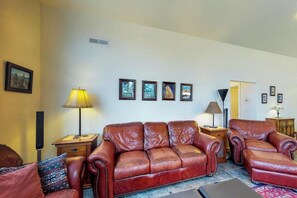 Ein Wohnzimmer mit braunem Ledersofa, einem passenden Sessel und einem kleinen Tisch mit einer Lampe. An der Wand befinden sich gerahmte Bilder und eine Tür zu einem anderen Raum.