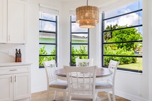 A kitchen with white cabinets, a dining table with four chairs, a hanging woven light fixture, and large windows with a view of greenery. - Gorgeous 4 BR, Pool Home, Game Room, Canal View, Sleeps 11! (Cape Coral)