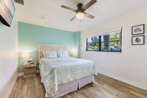 Ein Schlafzimmer mit Bett, Nachttisch, Deckenventilator, Fenster und Wandbildern.