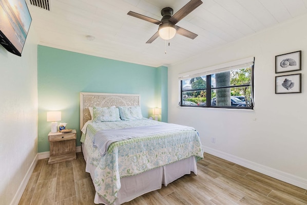 Un dormitorio con cama, mesita de noche, ventilador de techo, ventana y decoración en la pared.