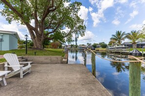 A waterfront patio with a concrete deck, a wooden chair, and a large tree. - Gorgeous 4 BR, Pool Home, Game Room, Canal View, Sleeps 11! (Cape Coral)