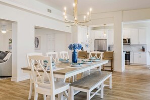 A modern kitchen with white cabinetry, a stainless steel refrigerator, and a dining area with a wooden table and white chairs. - Gorgeous 4 BR, Pool Home, Game Room, Canal View, Sleeps 11! (Cape Coral)